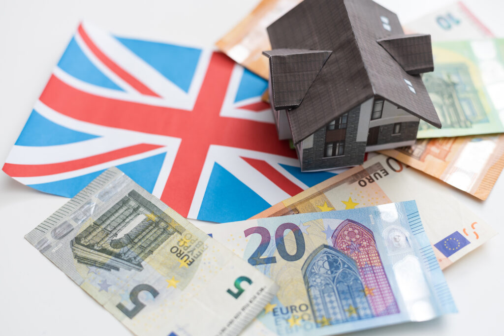 A model house placed on euro banknotes with a UK flag in the background, symbolizing housing and cost of living in the UK for migrants.