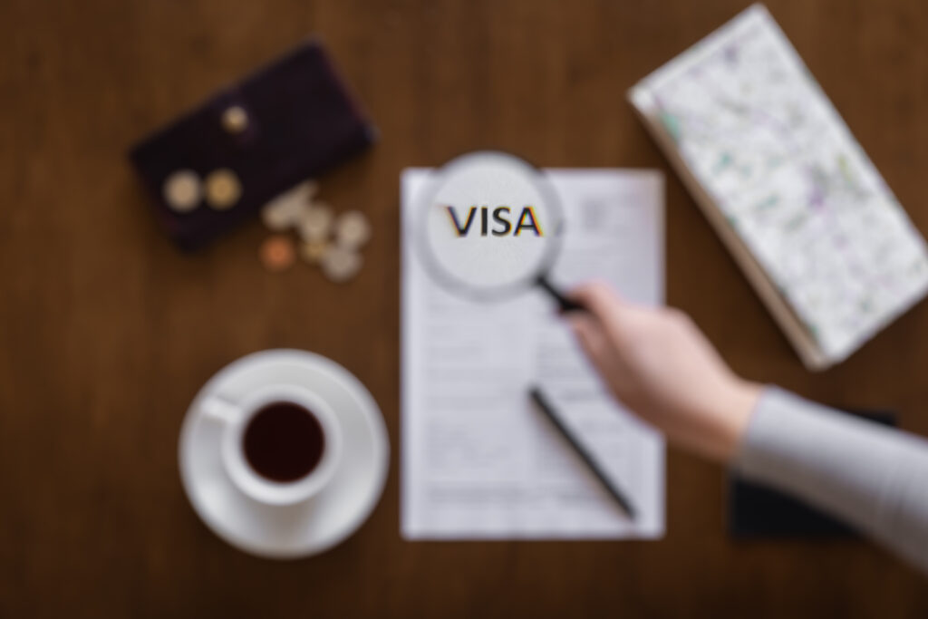 Magnifying glass highlighting the word “VISA” on a UK visa application form with coffee, pen, and wallet on a wooden table.