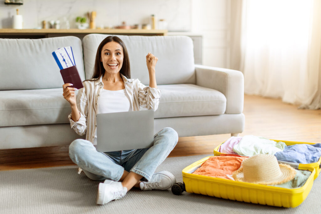 Smiling woman with passport and boarding passes using a laptop while packing luggage, symbolising new arrivals looking for accommodation in the UK.
